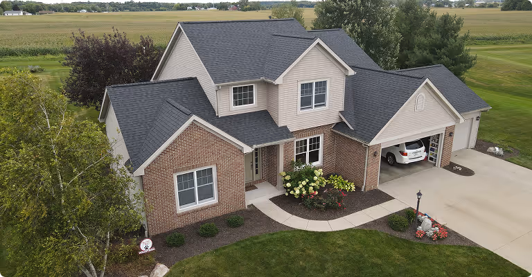 A large suburban house with a brick and siding exterior, a dark shingled roof, landscaped front yard, and a driveway leading to a two-car garage with a white car inside.