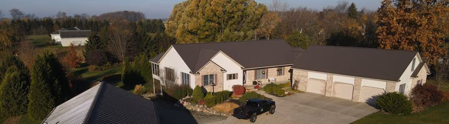 Aerial view of a single-story suburban house with beige brick exterior, a three-car garage, and a black pickup truck parked on the driveway, surrounded by autumn trees.