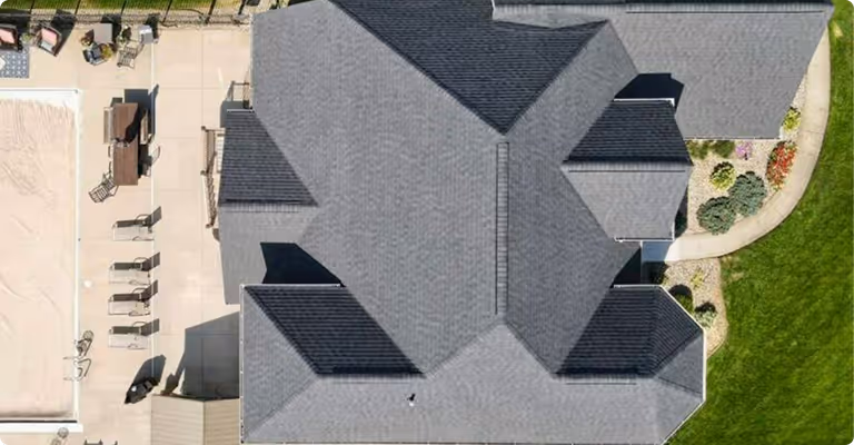 Aerial view of a large gray roofed building surrounded by patio furniture, swimming pool area, and green lawn with landscaped garden.