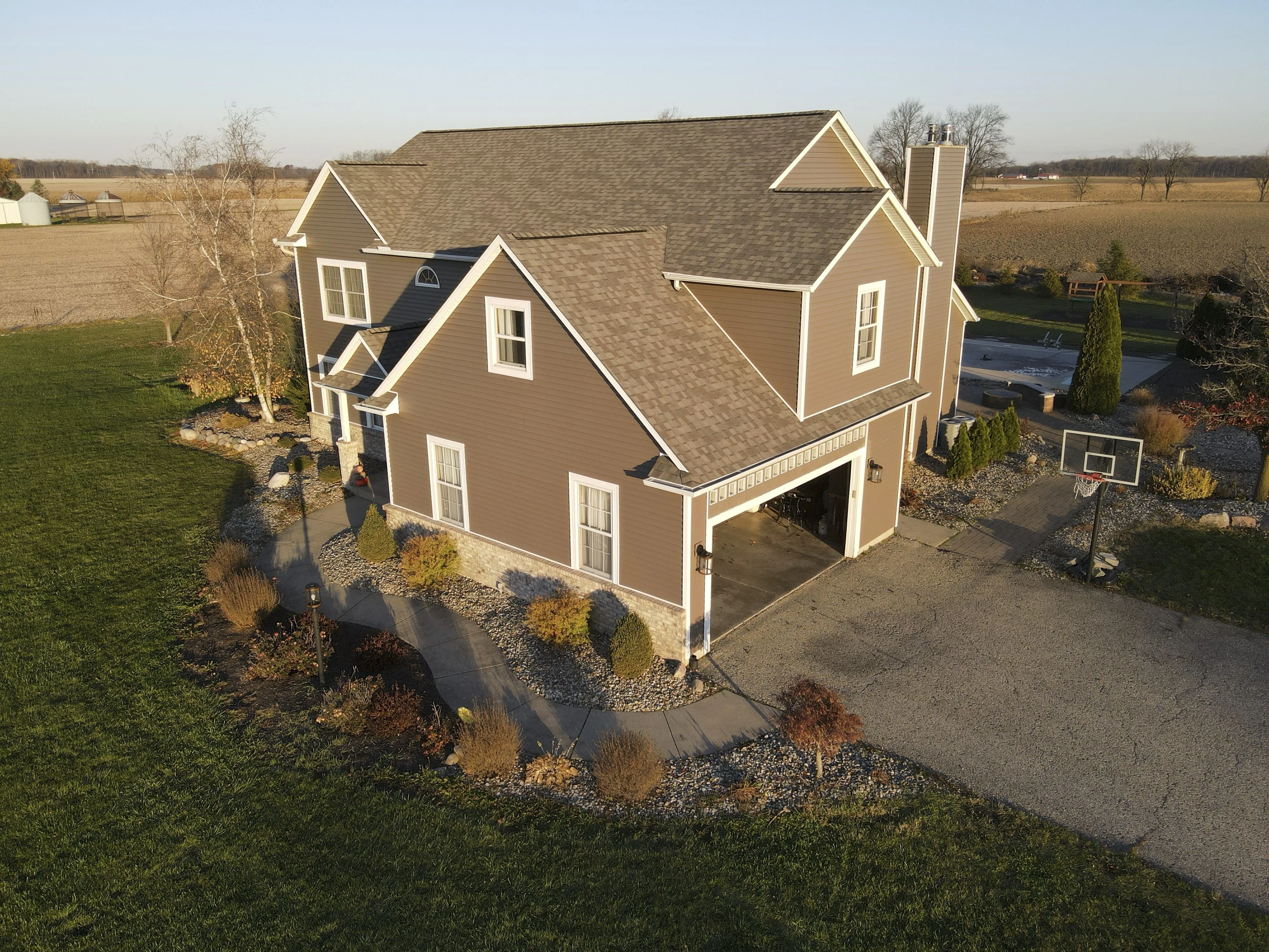 Two-story brown house with an open garage, curved walkway, landscaping, and a basketball hoop in a rural setting.