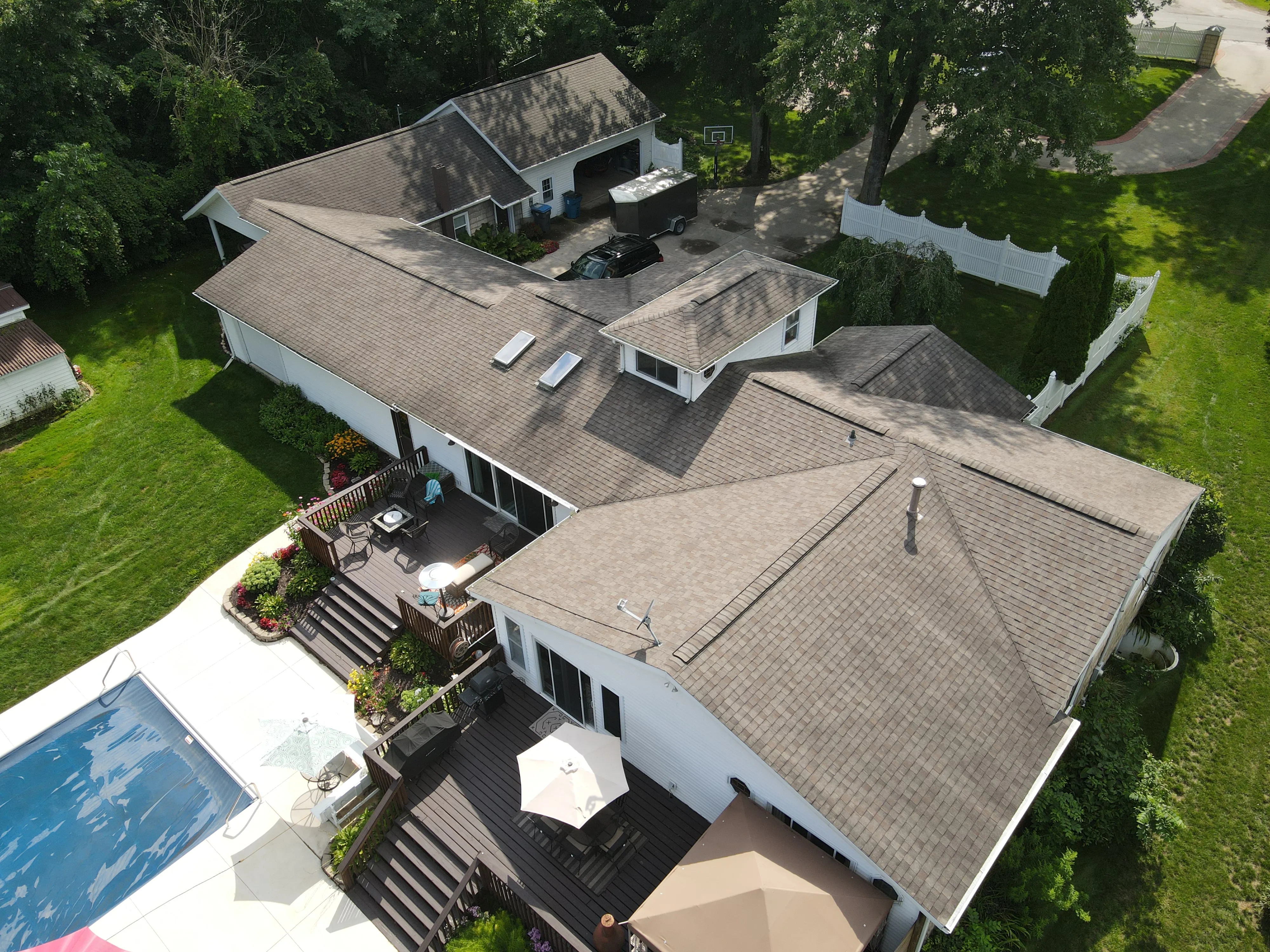 Aerial view of a large house with multiple roof sections, two wooden decks with umbrellas, stairs, and a swimming pool, surrounded by green lawn and trees.