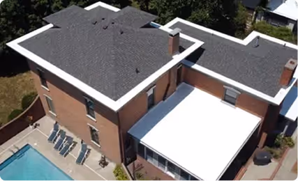 Aerial view of a large brick house with a dark shingle roof and a white flat roof section next to a swimming pool.