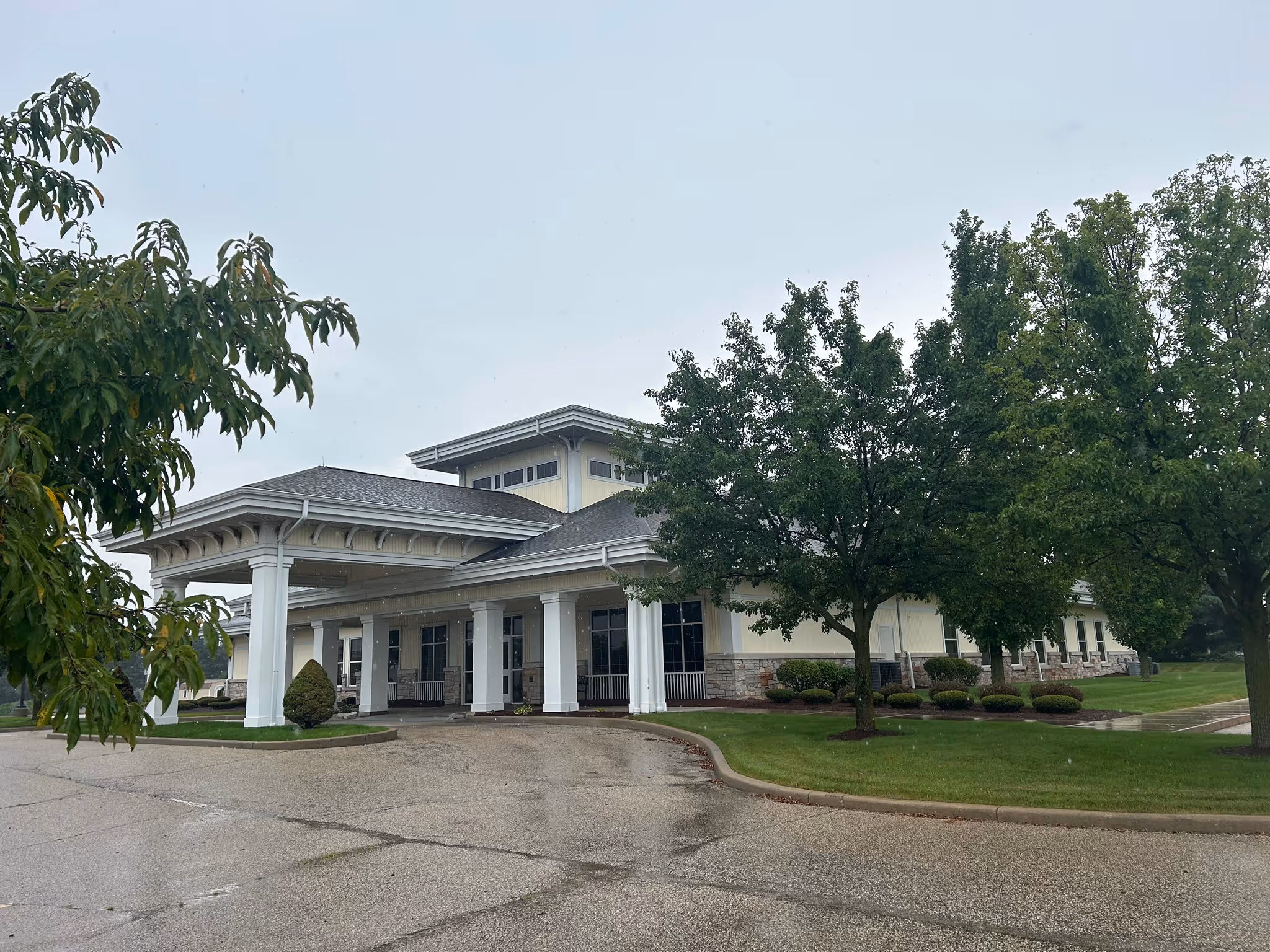 One-story building with a covered entrance, surrounded by green trees and a paved driveway on a cloudy day.