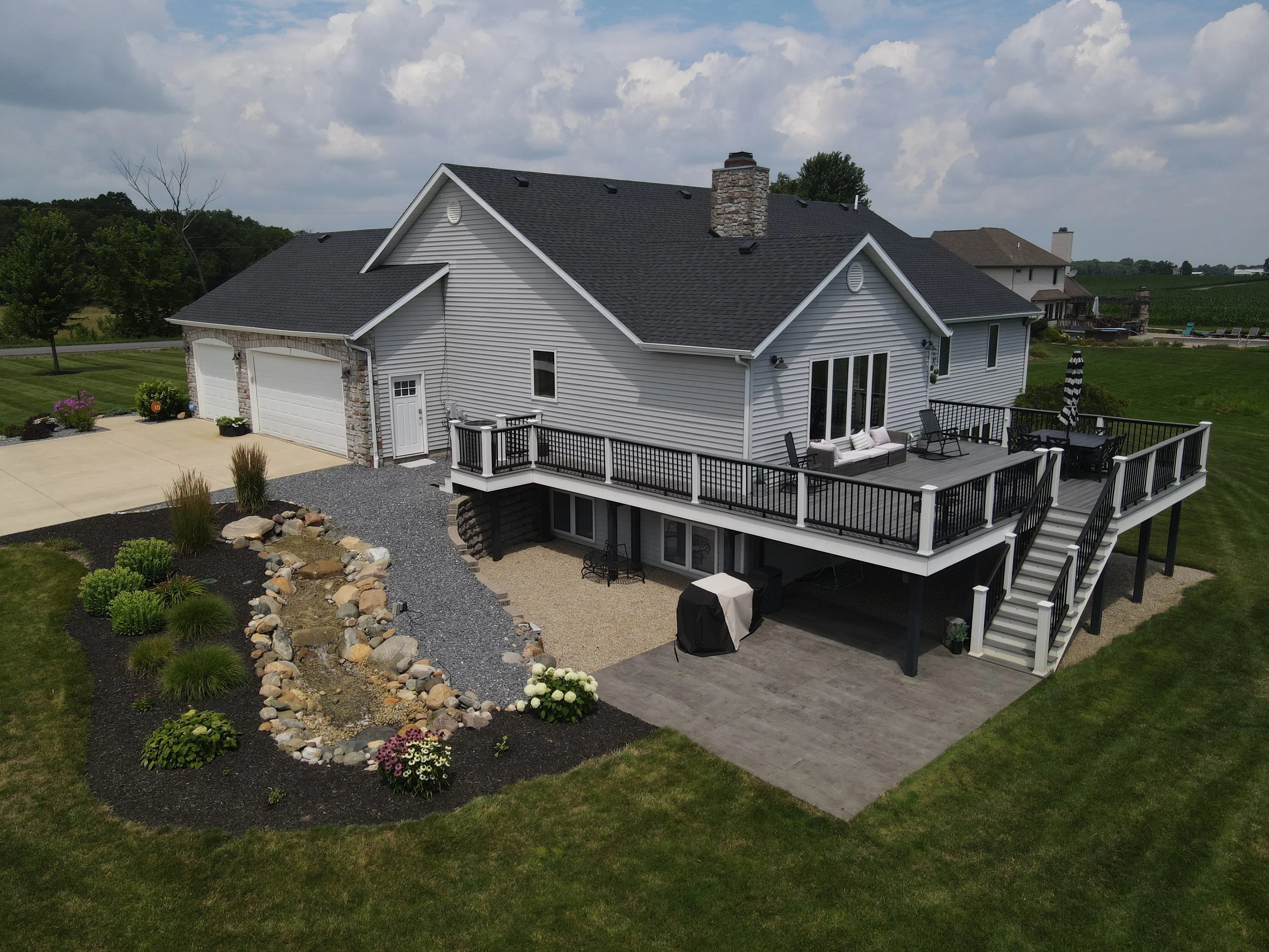 Modern house with gray siding, a large elevated deck with outdoor furniture, stairs leading to the lawn, landscaped garden with rocks and plants, and a three-car garage.