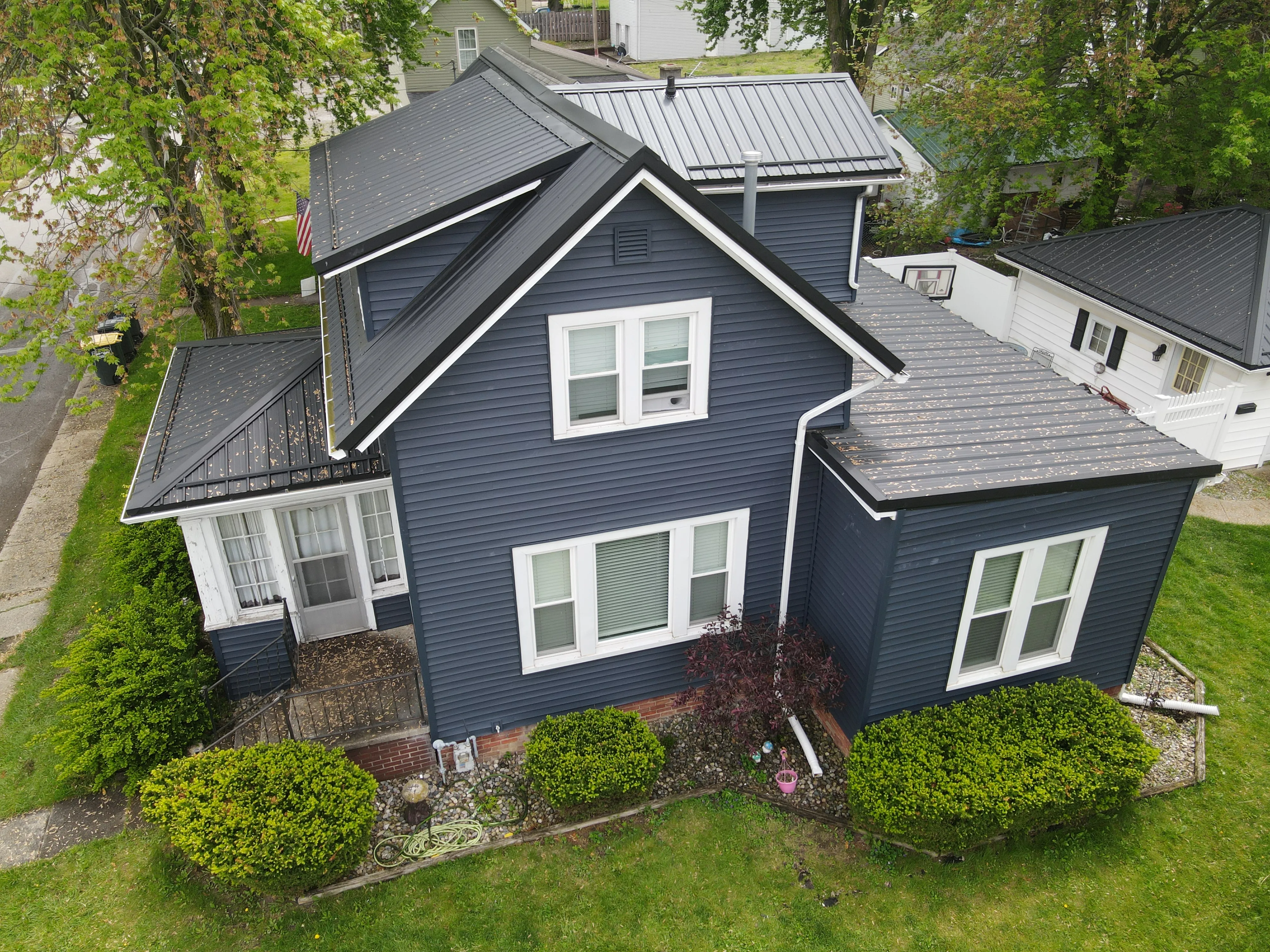 A two-story dark blue house with white window frames, a black metal roof, surrounded by green bushes and trees.