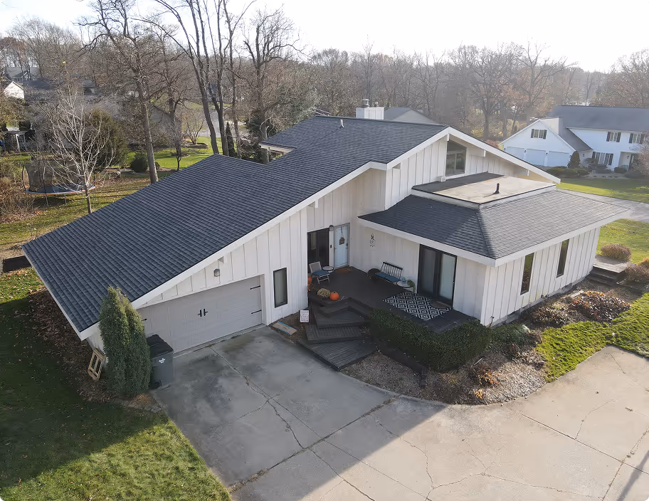 Modern single-story white house with multiple angled dark gray roofs, a garage, front porch with seating, and surrounding yard with trees and shrubs.