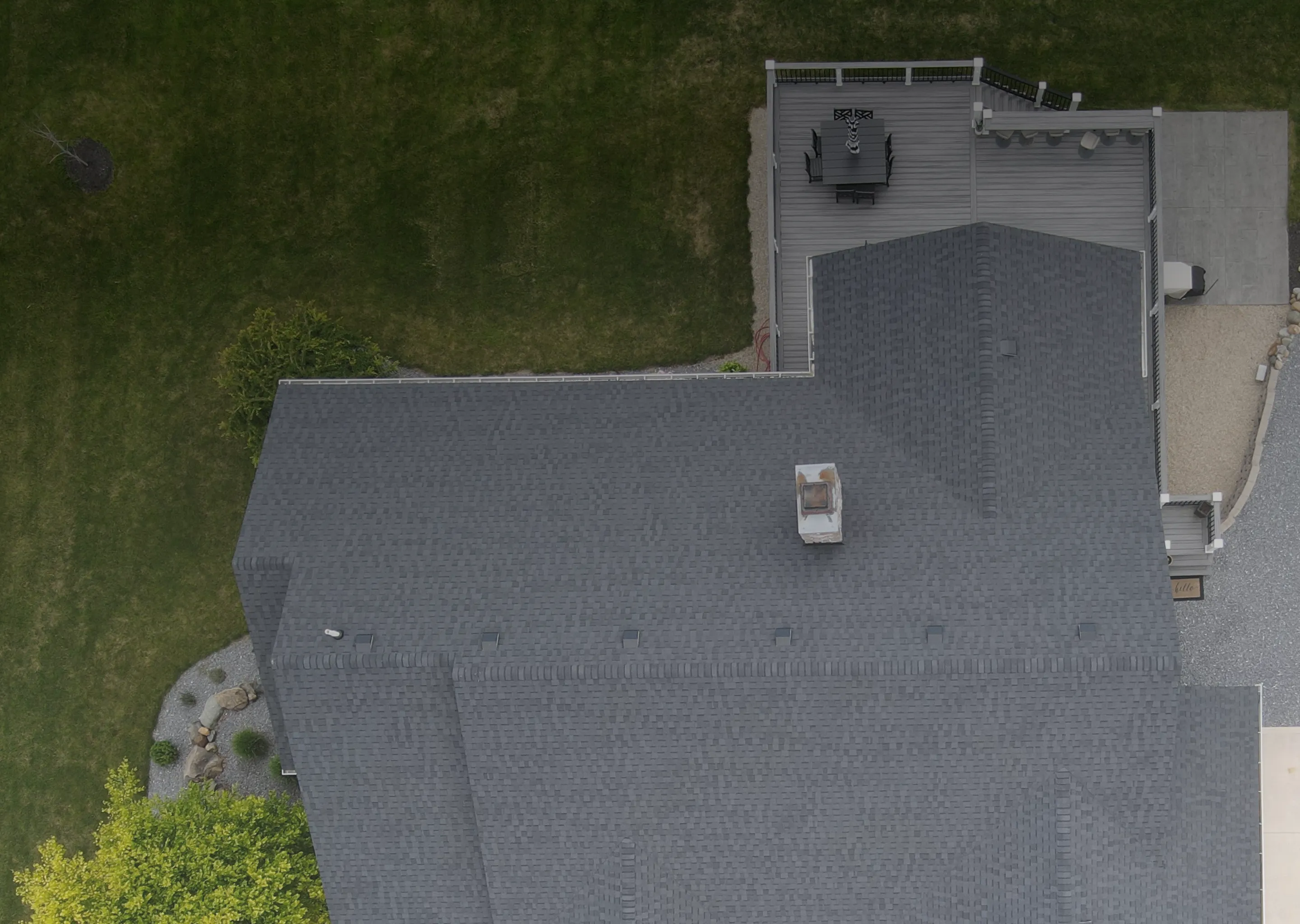 Aerial view of a dark gray shingled roof with a chimney and a deck with outdoor furniture, surrounded by green grass and trees.