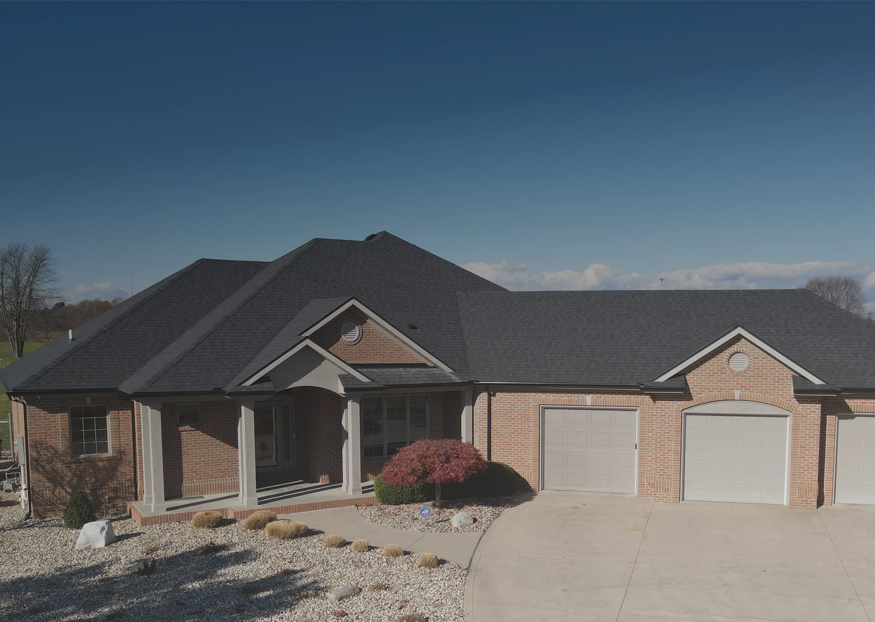 Single-story brick house with a dark roof, three garage doors, a small front porch, and a landscaped yard with gravel and bushes under a clear blue sky.