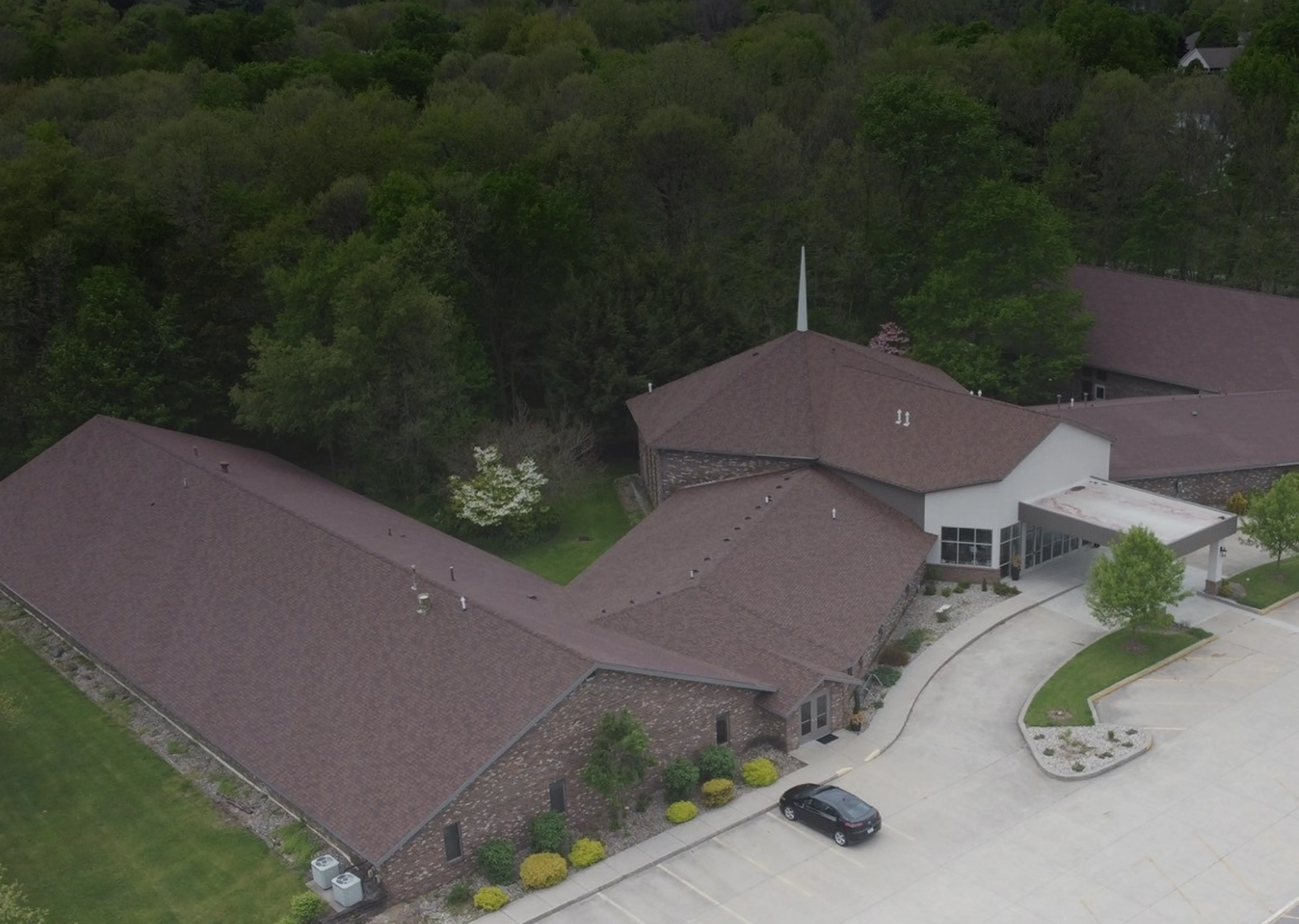 Aerial view of a large brick building with a brown roof, surrounded by trees and greenery, with a parking lot and a single black car parked near the entrance.