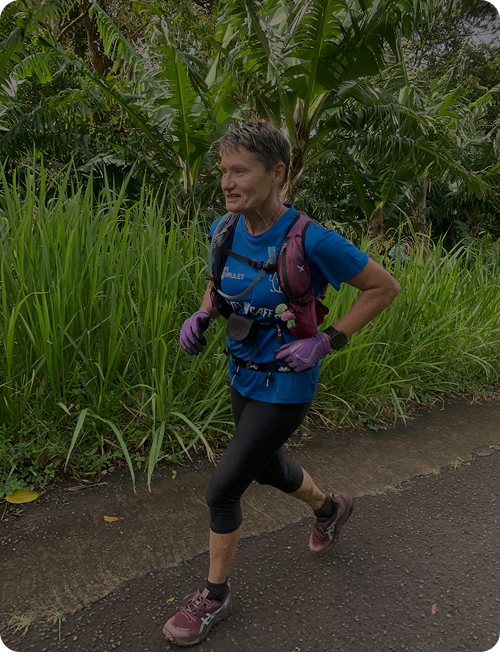 Femme courant sur une route bordée de végétation tropicale dense, portant un t-shirt bleu, des gants violets et un sac à dos de course.
