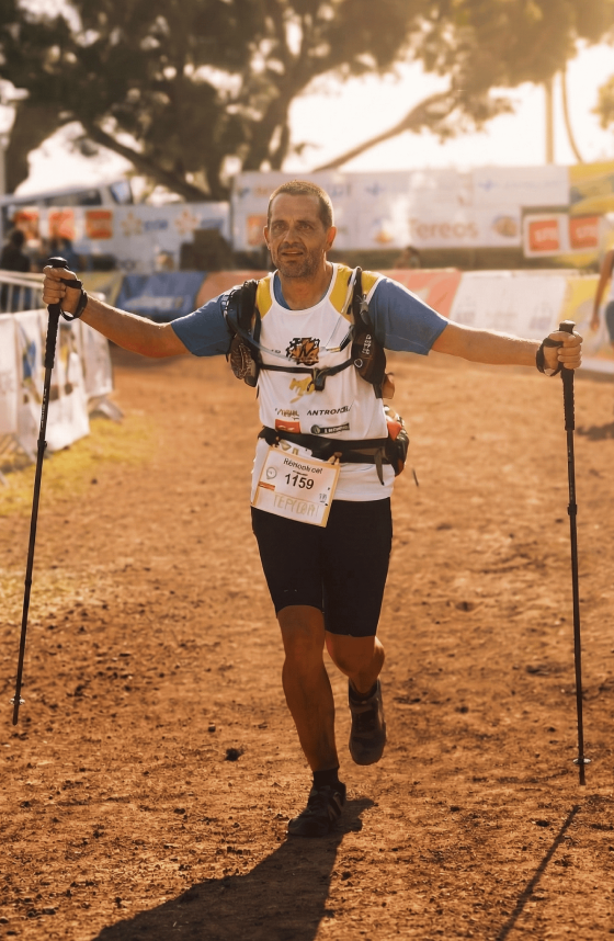 Un homme en tenue de course avec un dossard 1159 utilise des bâtons de randonnée sur un sentier en terre pendant une course en plein air au soleil couchant.