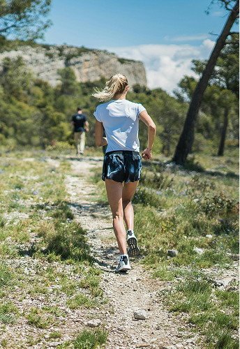Femme courant sur un sentier en pleine nature avec un homme devant elle, montagnes en arrière-plan.