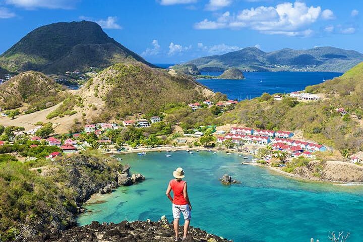 Personne debout au sommet d'une colline surplombant une baie turquoise avec des maisons colorées et des montagnes en arrière-plan.