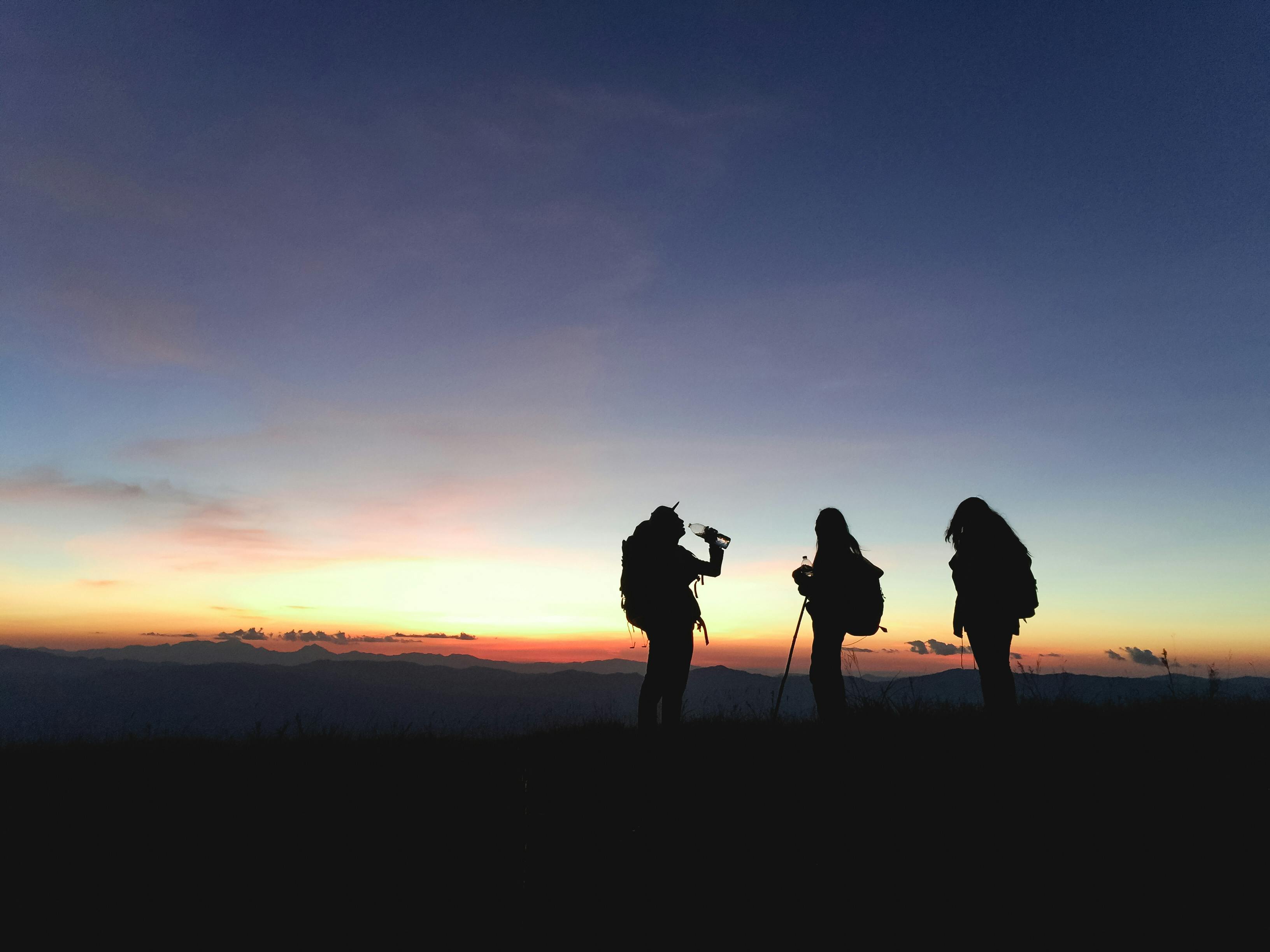 Silhouettes de trois randonneurs debout sur une colline au coucher du soleil avec un ciel coloré en arrière-plan.