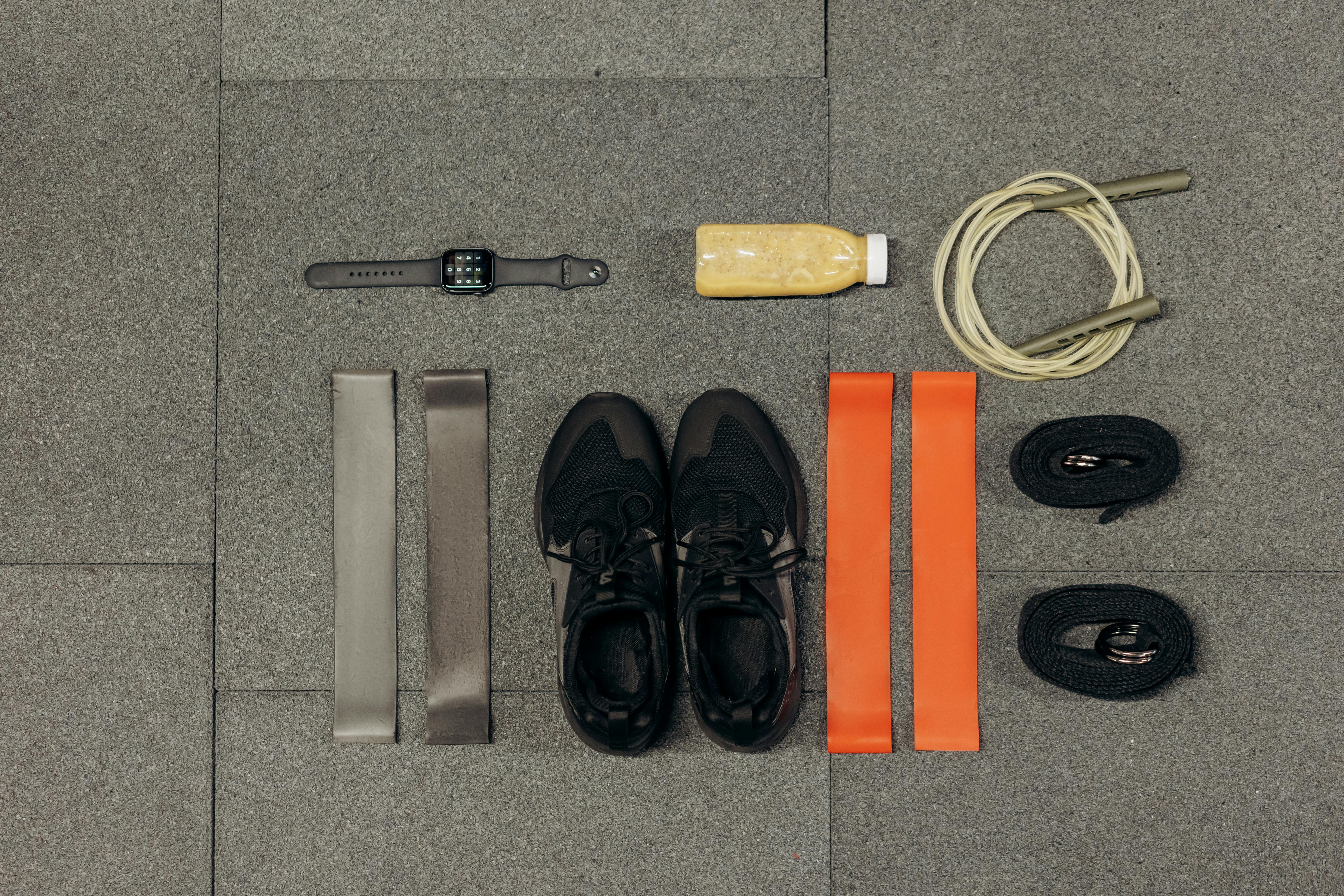 Équipement d'entraînement comprenant des bandes de résistance grises et orange, une paire de baskets noires, une corde à sauter beige, une montre connectée noire, et une bouteille de boisson protéinée beige disposés sur un sol gris.