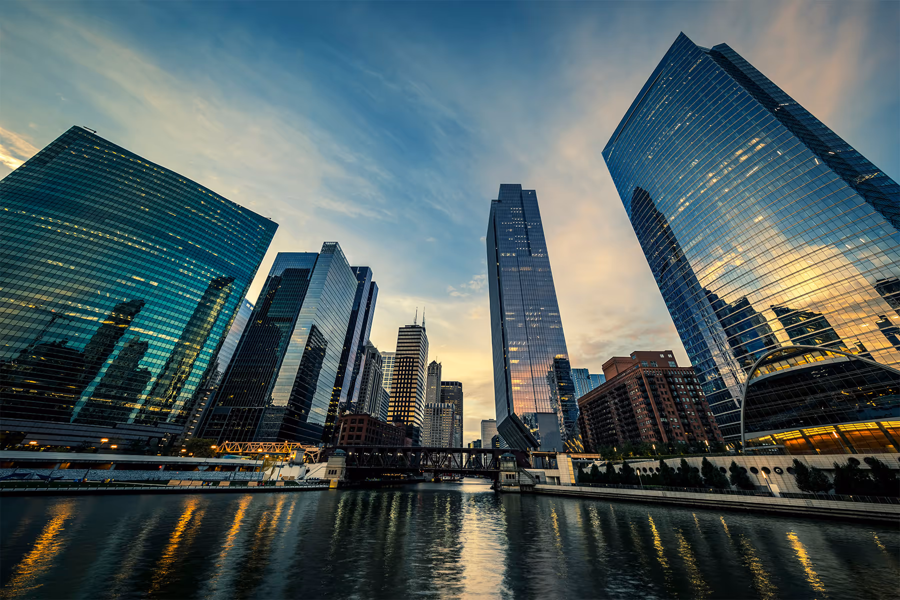Sunset view of a city skyline with modern glass skyscrapers reflecting light over a river with a bridge crossing it.