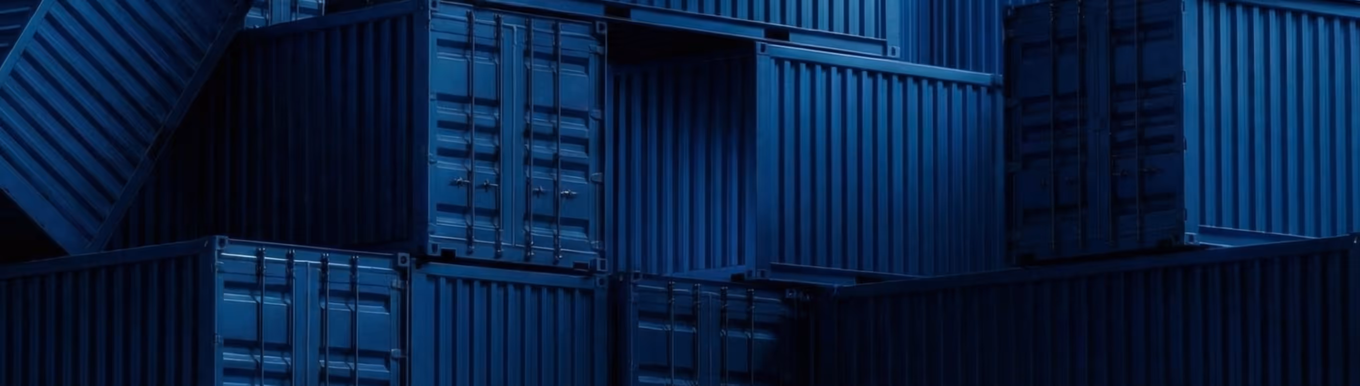 Stacked dark blue shipping containers in dim light.