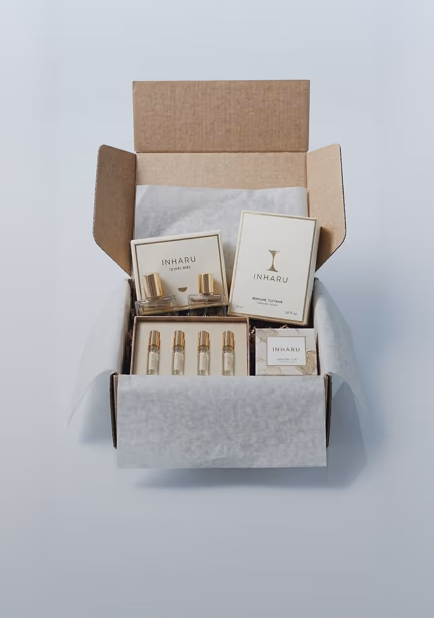Open cardboard box with Inharu perfume products including two full-size bottles, four small vials, and product boxes on white tissue paper.