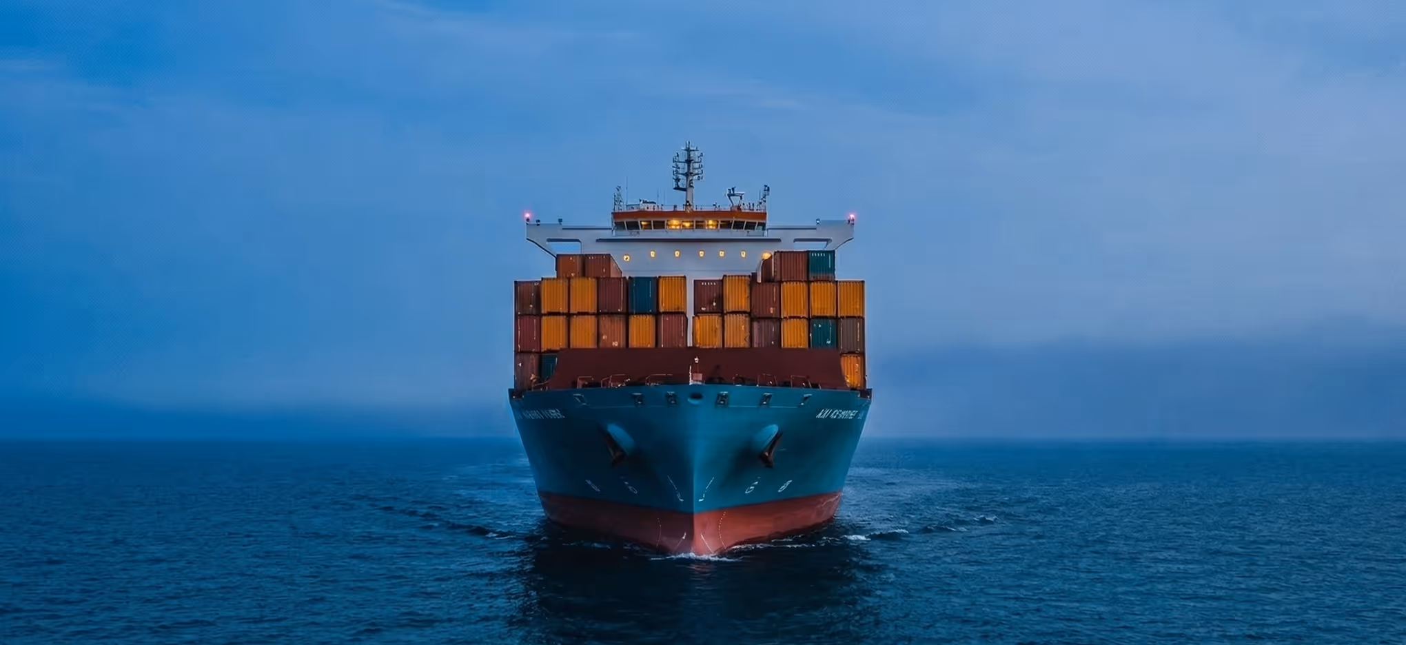 Front view of a large container ship loaded with colorful shipping containers sailing on calm blue ocean under a clear sky.