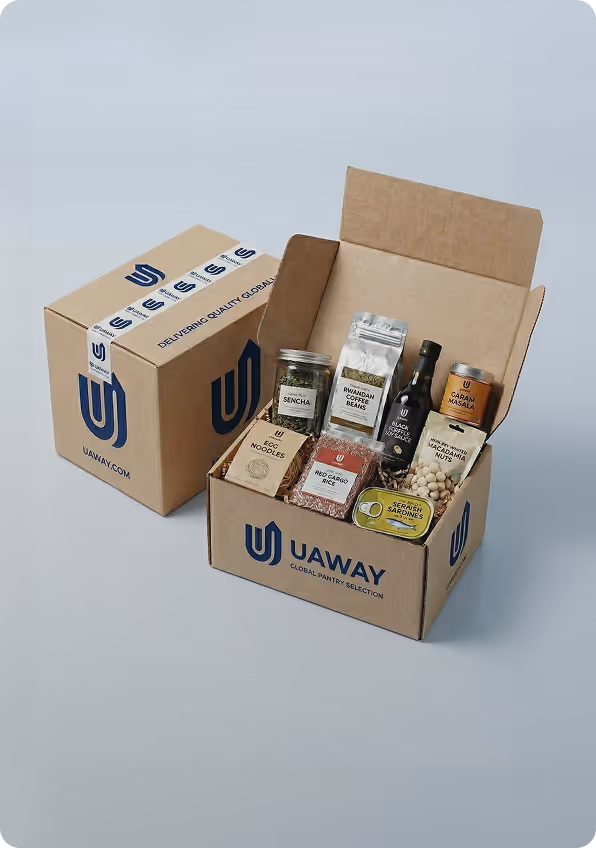 Open Uaway cardboard box containing packaged food items like sencha tea, Rwandan coffee beans, black truffle oil, garam masala, egg noodles, red cargo rice, smoked macadamia nuts, and sardines, with a closed similar box beside it.