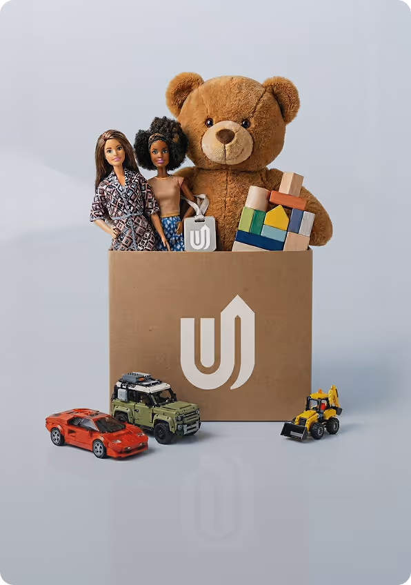 Cardboard box with a teddy bear, two dolls, and colorful building blocks inside, surrounded by three toy vehicles.