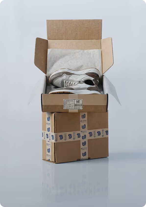 Pair of gray and white sneakers inside an open cardboard shoebox, placed on top of a sealed cardboard box with branded tape.