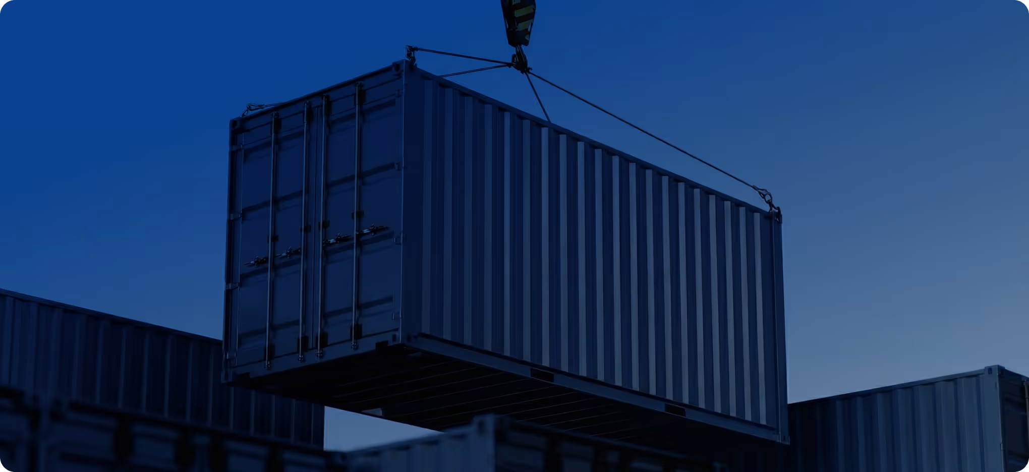 Shipping container being lifted by crane at dusk over other containers.