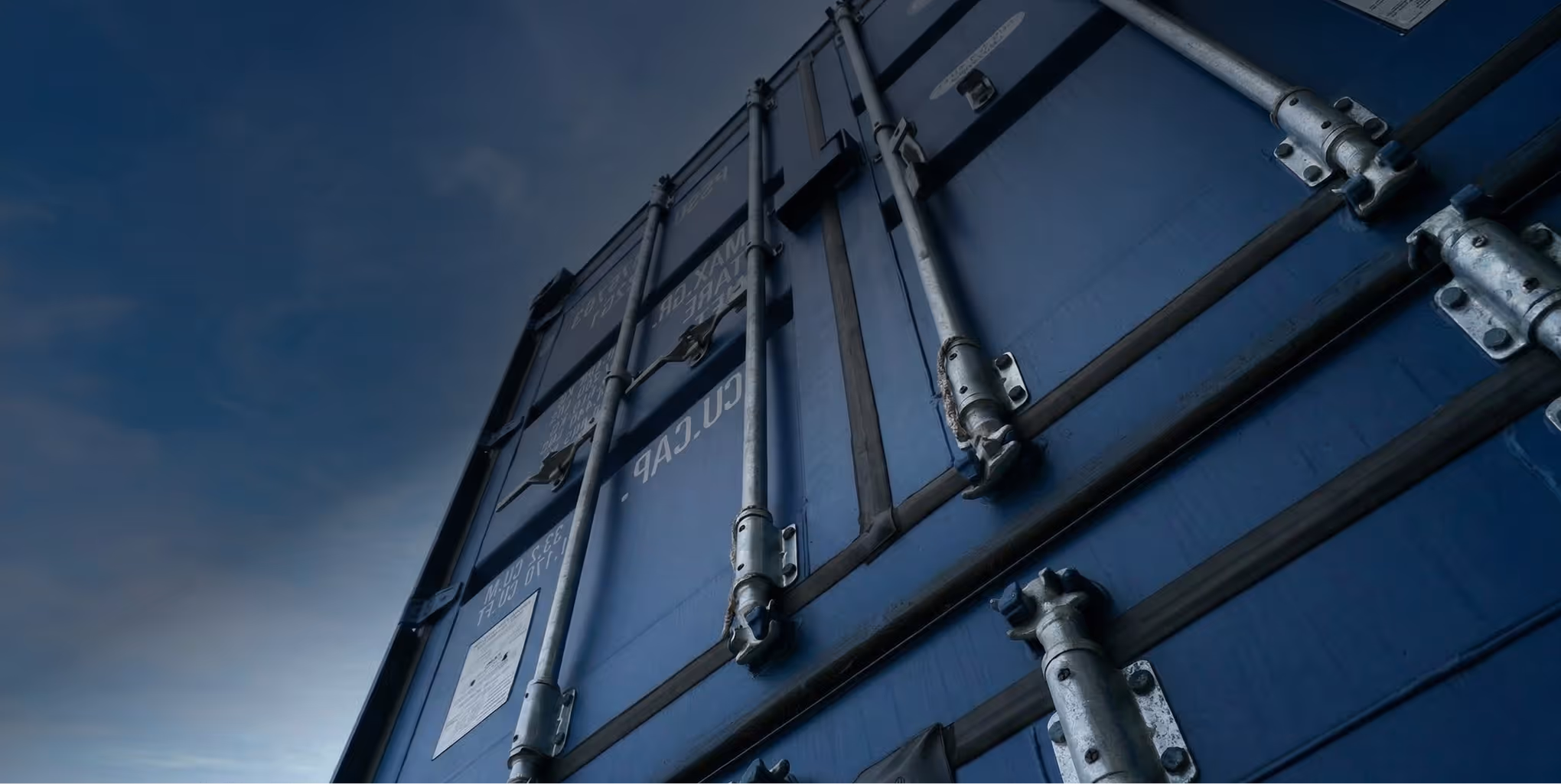 Close-up view of a closed blue shipping container door with metal locking bars against a cloudy sky.