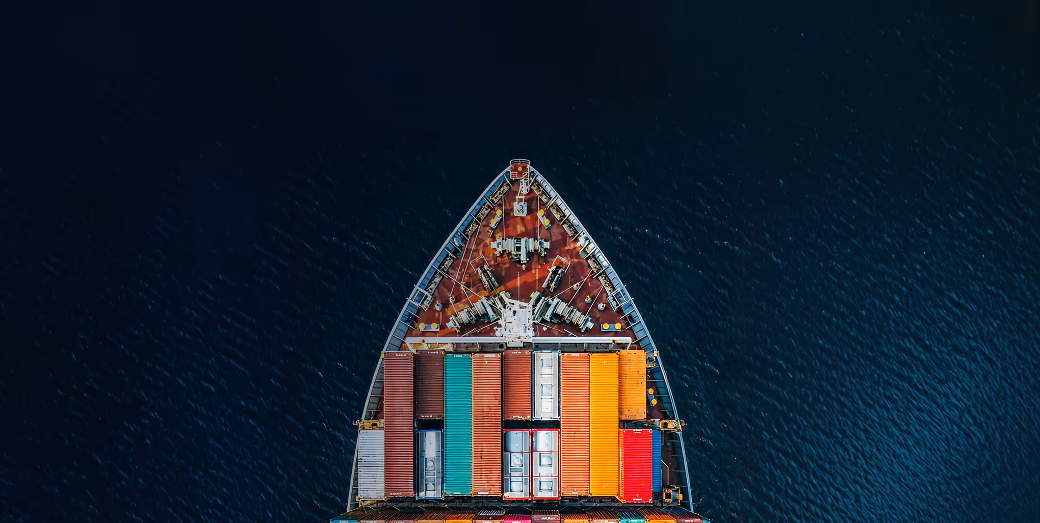 Aerial view of the bow of a cargo ship loaded with colorful shipping containers on dark blue water.