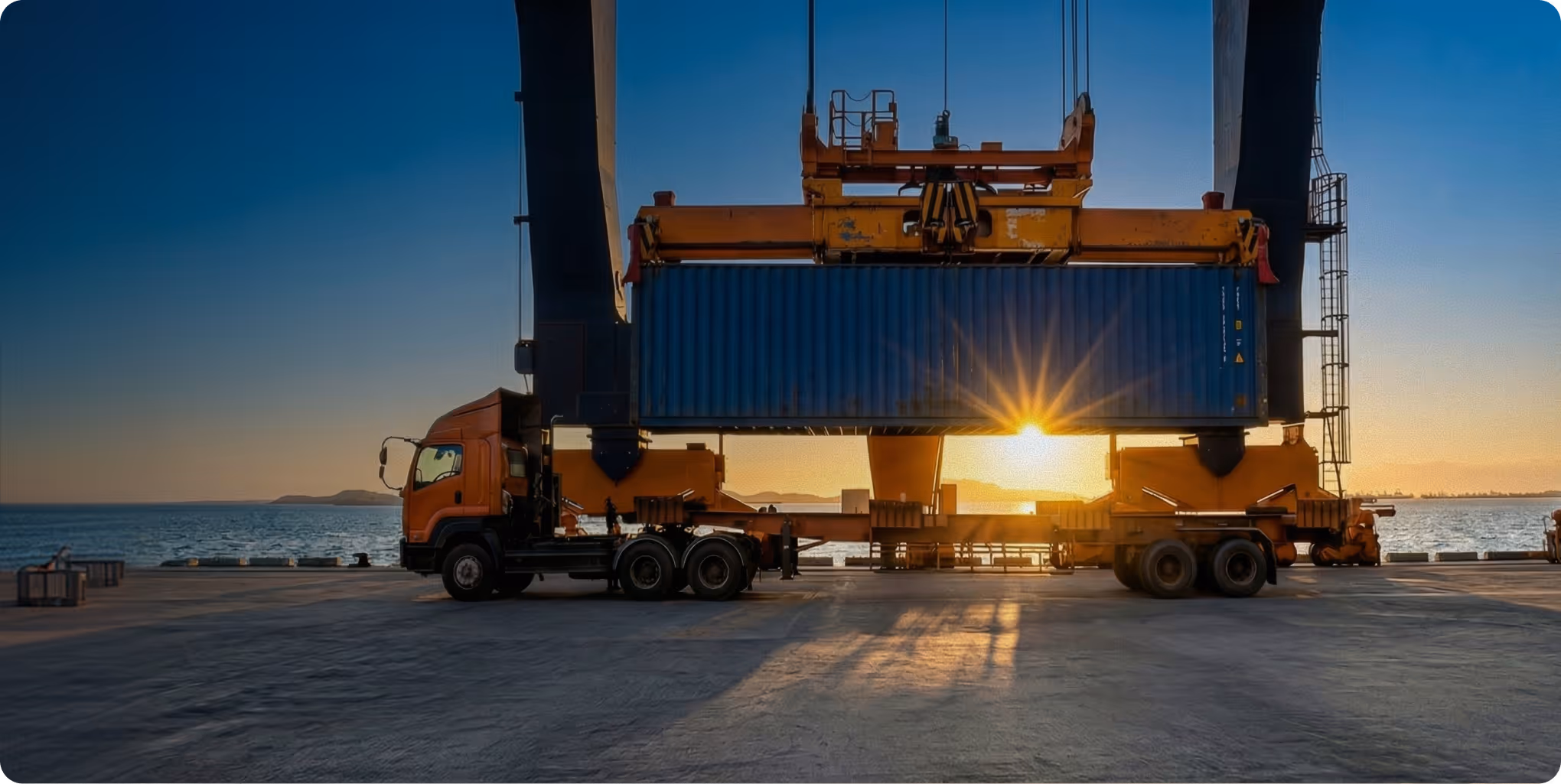 Orange truck with a blue cargo container being lifted by a crane at a port during sunset over the sea.
