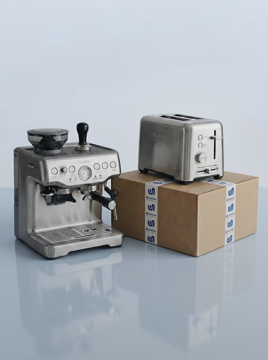 Stainless steel Breville espresso machine next to a Breville toaster placed on a cardboard box sealed with Uaway tape on a reflective surface.