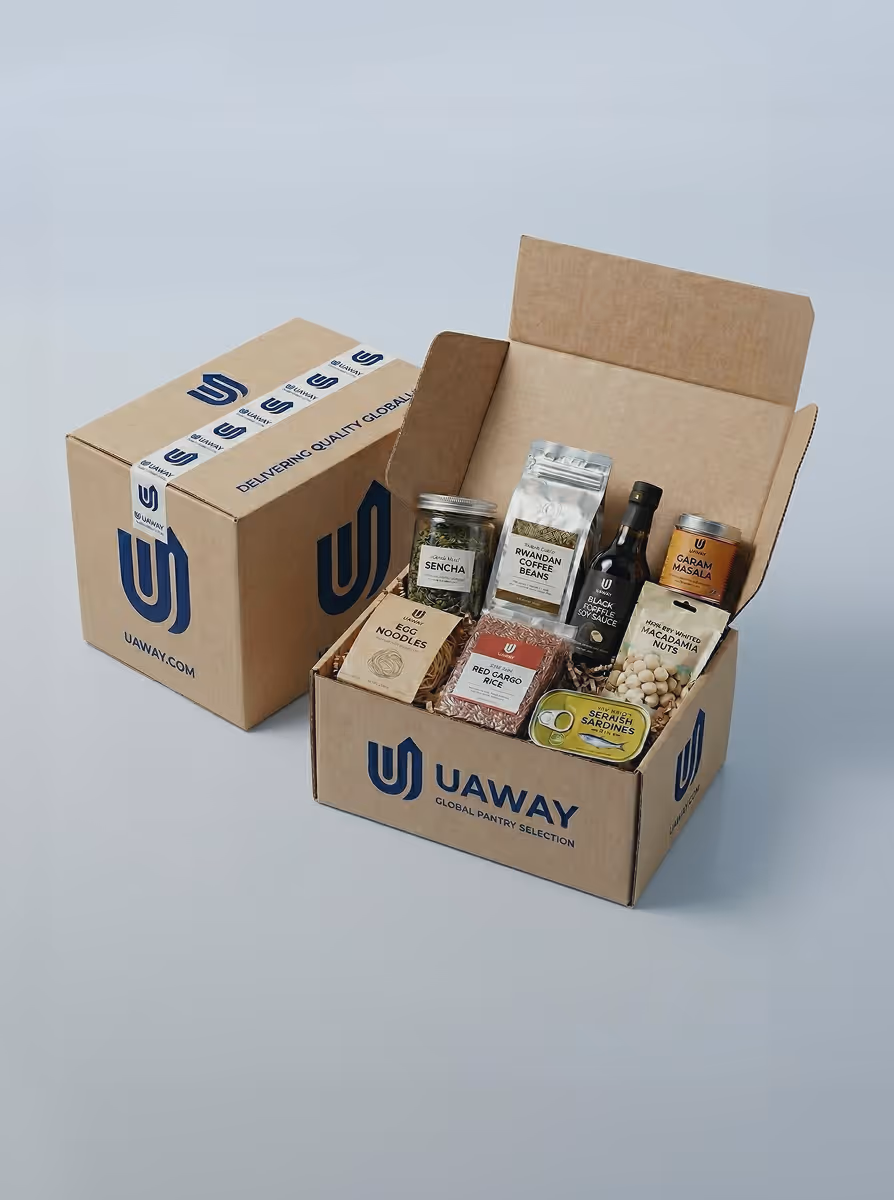 Open cardboard box labeled UAWAY containing various pantry items including Sencha tea, Rwandan coffee beans, black coffee soy sauce, garam masala, egg noodles, red cargo rice, serarish sardines, and macadamia nuts.