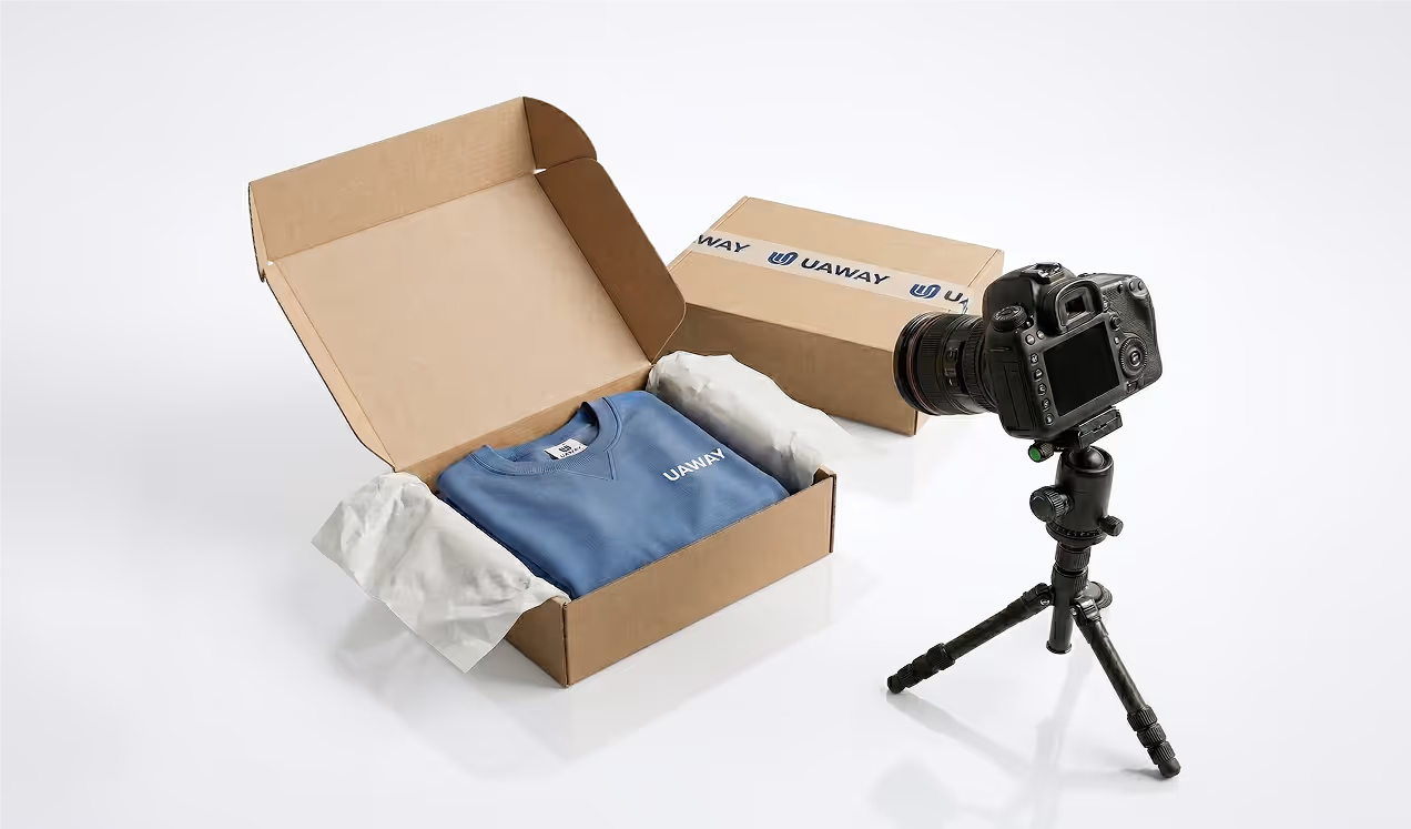 Open cardboard box containing a folded blue sweatshirt with 'UAWAY' logo, next to a closed box and a DSLR camera on a tripod.