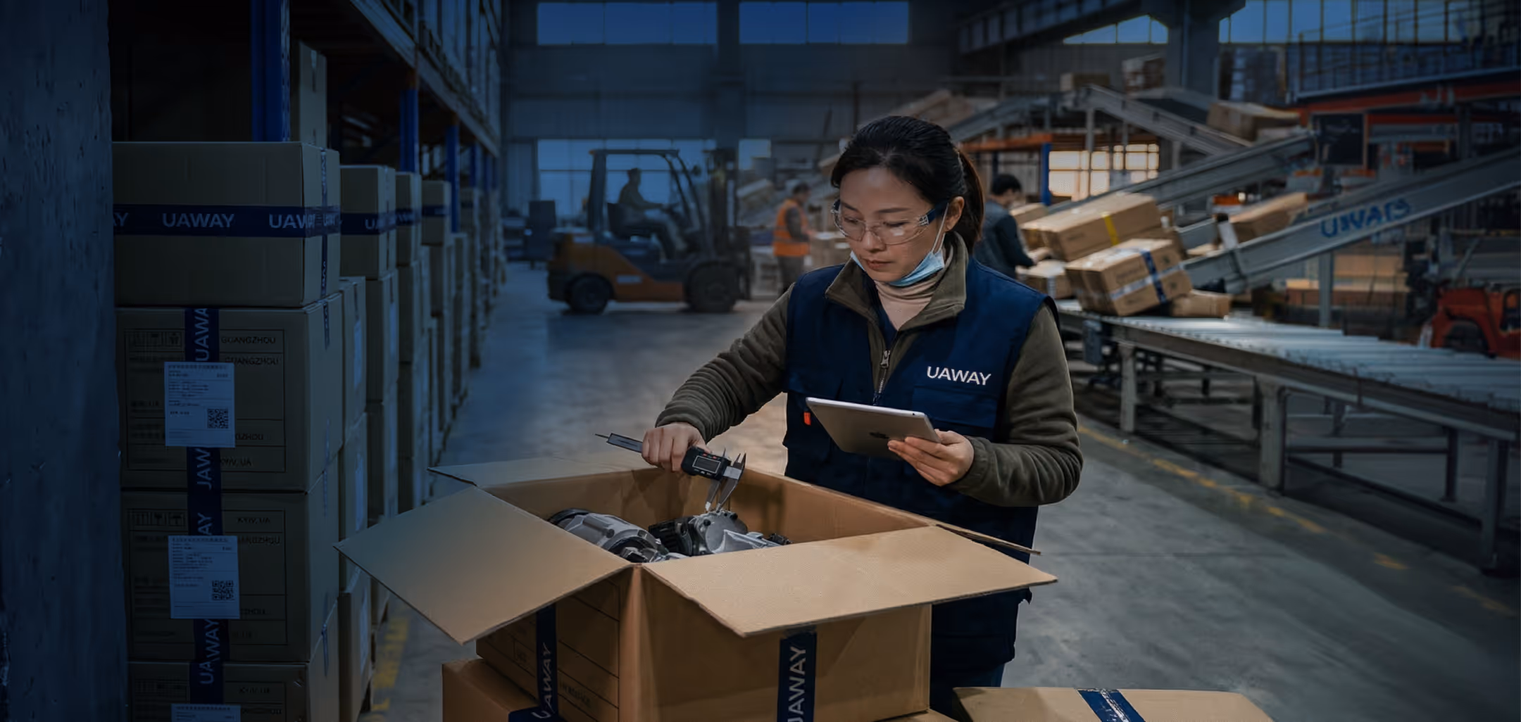 Warehouse worker measuring parts inside a large cardboard box using calipers while holding a tablet in an industrial setting.