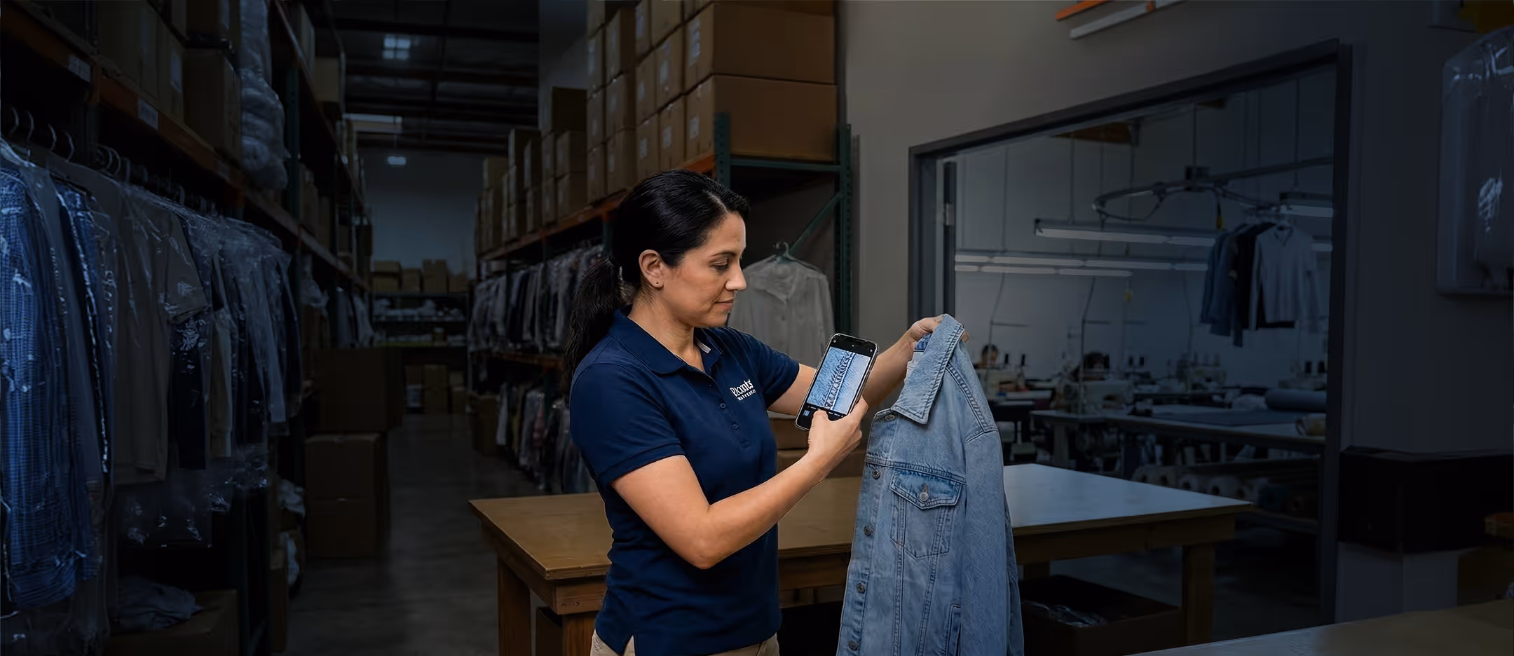 Woman in a warehouse inspecting a denim jacket and taking a close-up photo of its fabric with a smartphone.