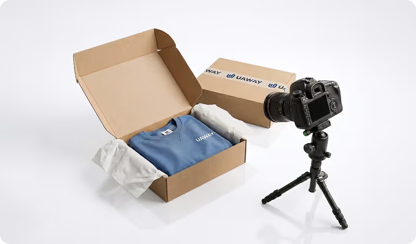 Blue UAWAY branded t-shirt neatly folded in an open cardboard box with tissue paper, next to a professional camera on a tripod.