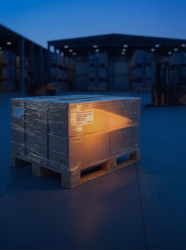 Stack of cardboard boxes wrapped in plastic on a wooden pallet in a dimly lit warehouse with a warm light shining on the boxes.