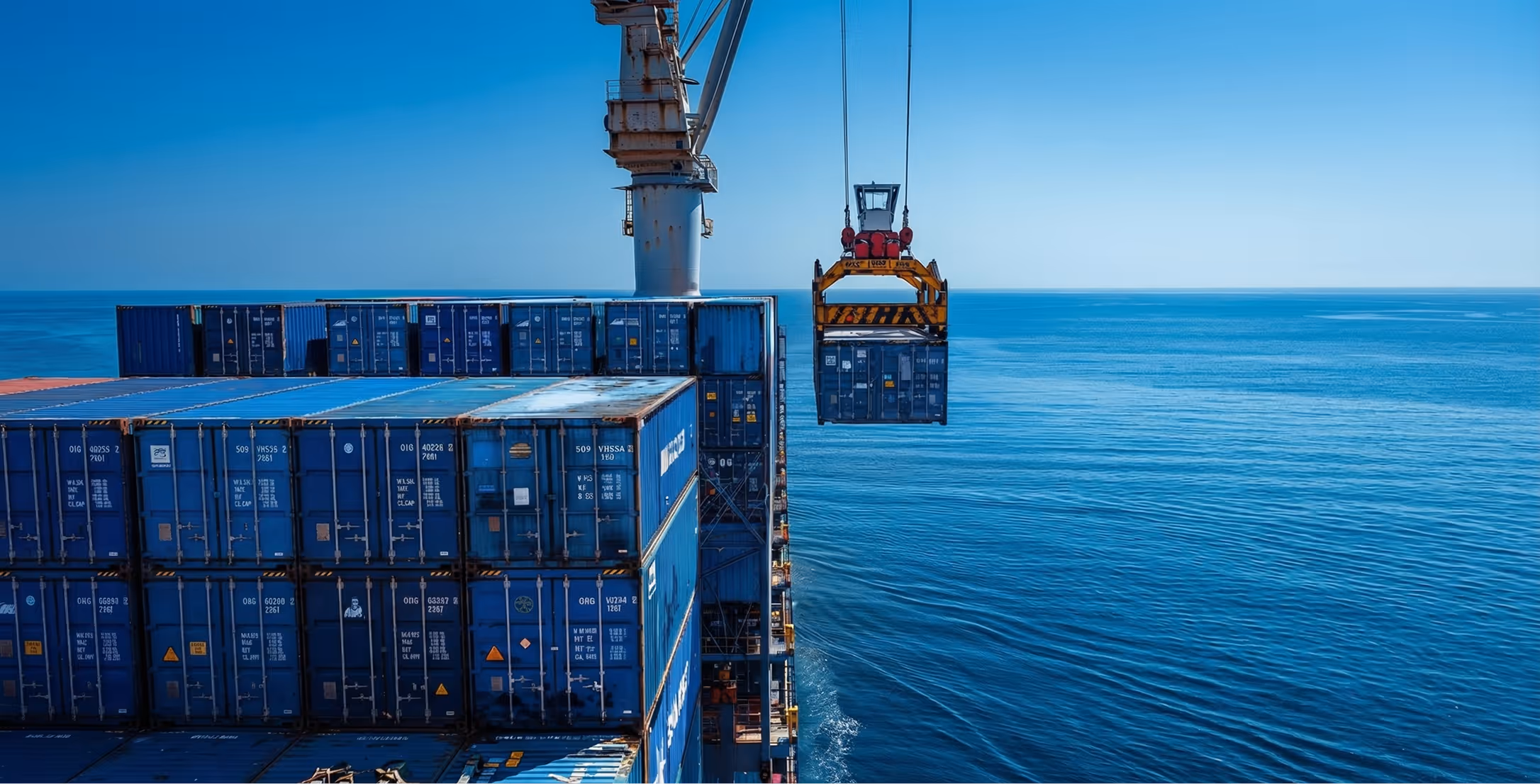 Crane lifting a blue shipping container on a cargo ship at sea with stacked blue containers and clear sky.