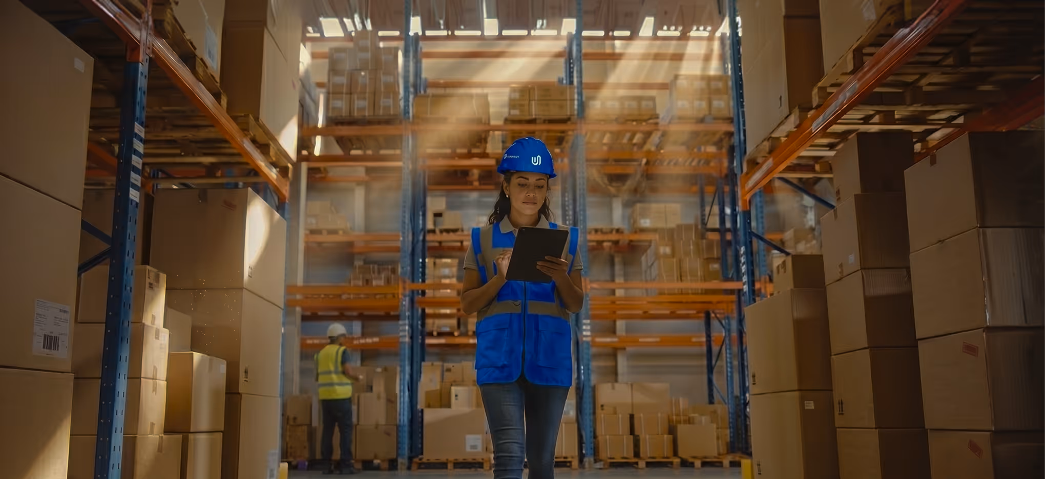 Woman in a blue safety vest and hard hat using a tablet in a warehouse with shelves full of boxes.