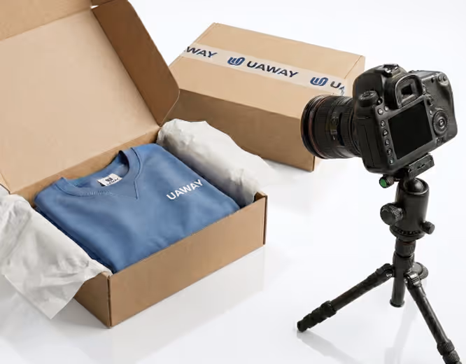Blue UAWAY branded sweatshirt neatly folded inside an open cardboard box with tissue paper, next to a camera on a tripod.