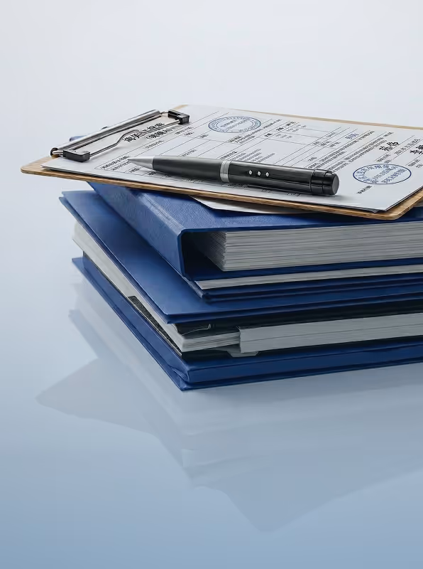 Stack of blue binders topped with a clipboard holding a document and a black pen on a reflective surface.