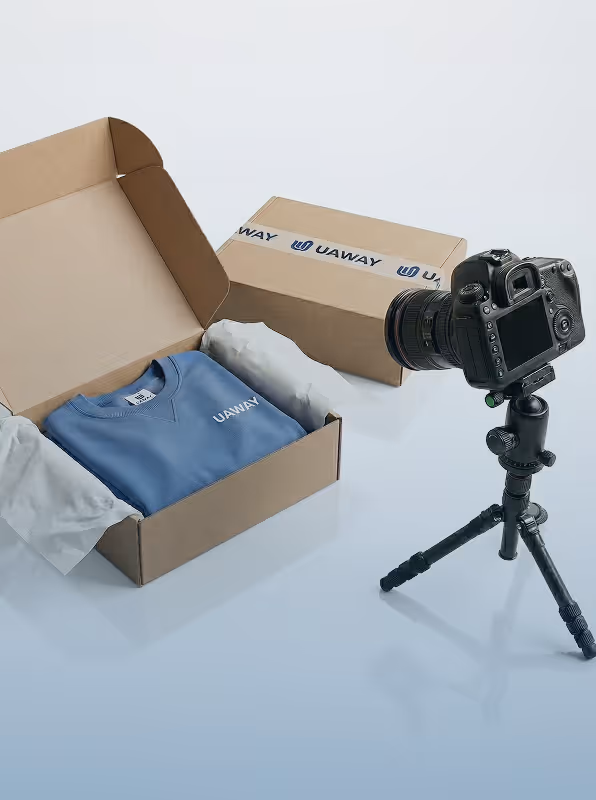 Product photography setup with a camera on a tripod facing a cardboard box containing a blue sweatshirt labeled 'UAWAY', and another sealed box in the background.