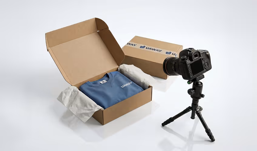 Open cardboard box with a blue UA Way sweatshirt inside and a camera on a tripod positioned to photograph the box.