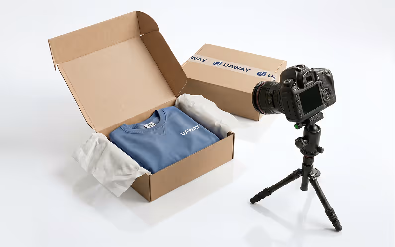 Open cardboard box with folded blue sweatshirt labeled UAWAY and white tissue paper, next to a packaged box and a DSLR camera on a tripod.