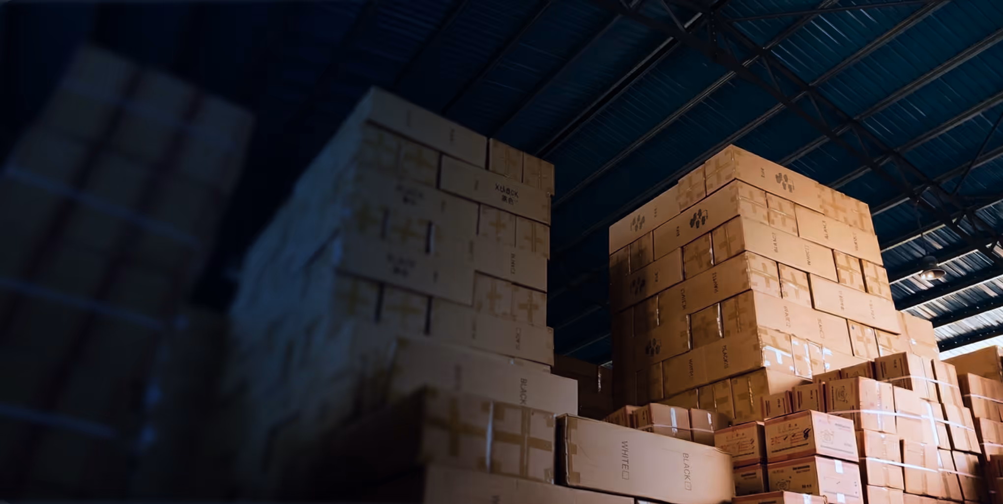 Stacks of cardboard boxes arranged in a warehouse with a metal roof overhead.