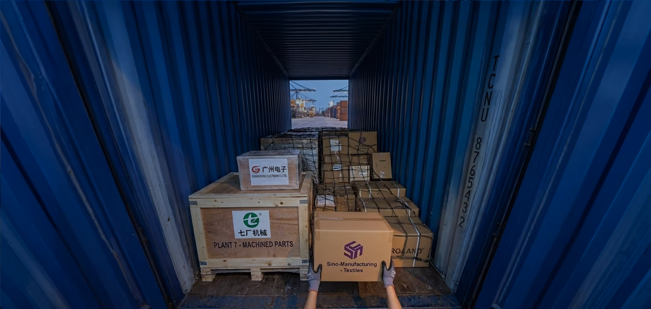 View from inside a blue shipping container filled with wooden crates and cardboard boxes labeled for machinery and textiles, hands holding a box labeled Sino-Manufacturing Textiles.
