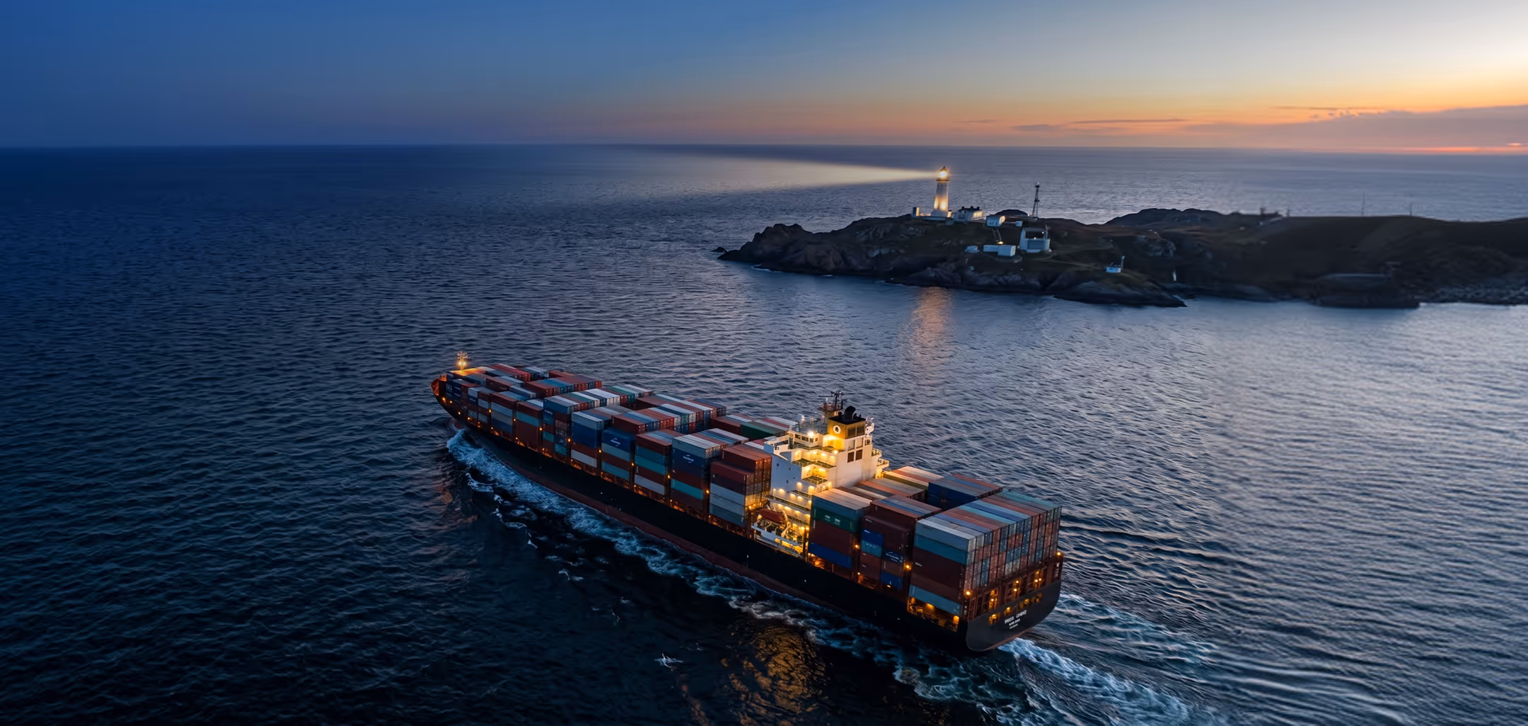 Large container ship sailing near a rocky island with a lighthouse at sunset.
