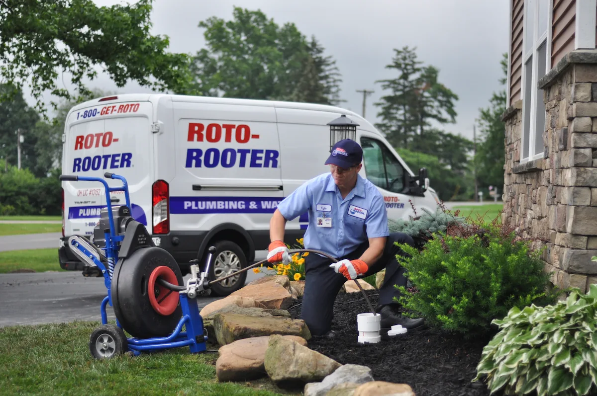 Roto-Rooter plumber using equipment to clear an underground pipe near a house with a Roto-Rooter van parked in the background.