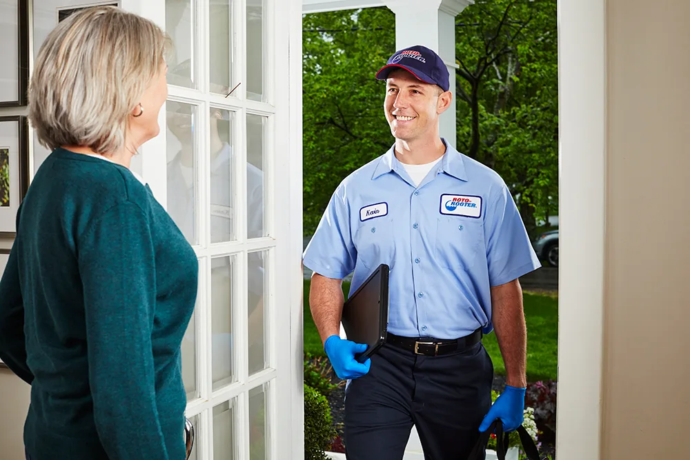 A Roto-Rooter technician wearing a blue uniform and gloves holds equipment and smiles at a woman standing in her doorway.