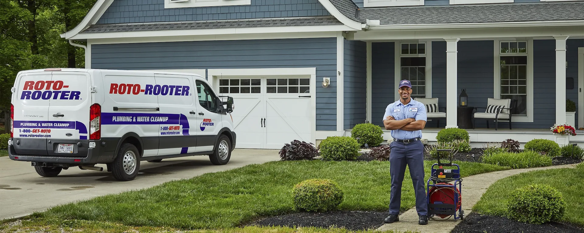 A Roto-Rooter technician standing on a walkway with plumbing equipment in front of a blue house and a Roto-Rooter service van parked in the driveway.